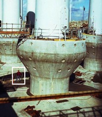 Základy pro gravitační uložení větrných elektráren na dno průlivu Middelgrunden v suchém bazénu, kde byly vyráběny. Na detailním záběru je možné porovnání jejich velikosti s postavou (v bílém rámečku). Foto B. Koč